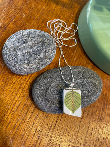 A BUTIK KUBIK Saved & Remade necklace Porcelain, featuring a green leaf design on a silver rectangular pendant, rests on gray stones atop wood, next to a round green object.