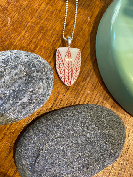 The Saved & Remade Necklace Porcelain by BUTIK KUBIK, featuring a red leaf motif on upcycled porcelain and hung on a silver chain, is displayed on wood with two gray stones and a green curved accent.
