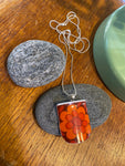 The Saved & Remade Necklace Porcelain by BUTIK KUBIK is a silverplated chain with a rectangular pendant made from recycled porcelain, showing a vivid orange and red flower. Displayed on gray stones, a green accent appears in the background.