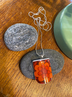 The Saved & Remade Necklace Porcelain by BUTIK KUBIK is a silverplated chain with a rectangular pendant made from recycled porcelain, showing a vivid orange and red flower. Displayed on gray stones, a green accent appears in the background.