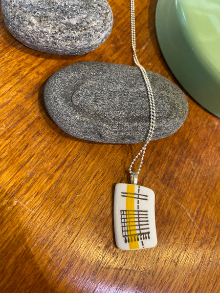 The BUTIK KUBIK Saved & Remade Necklace Porcelain features a yellow and black geometric pendant made from återbrukat porslin, shown on a smooth gray stone beside wood and green ceramic, hanging from a silverpläterad kedja.