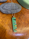 The BUTIK KUBIK Saved & remade necklace Porcelain features a rectangular green pendant of recycled porcelain with a black, white, and yellow geometric design. It is displayed on wood beside smooth gray stones and a green item.