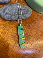 The BUTIK KUBIK Saved & remade necklace Porcelain features a rectangular green pendant of recycled porcelain with a black, white, and yellow geometric design. It is displayed on wood beside smooth gray stones and a green item.