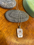 The Saved & remade necklace Porcelain by BUTIK KUBIK features a rectangular pendant with a delicate dandelion motif on återbrukat porslin, displayed on a smooth gray stone. A silverpläterad kedja completes its elegant look.
