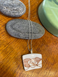 BUTIK KUBIKs Saved & remade necklace Porcelain: ett silverpläterat kedjehalsband med vitt återbrukat porslin och rött rosmotiv ligger på grå stenar på trä bredvid ett grönt föremål.
