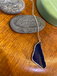 A silverplated chain with a blue, leaf-patterned Saved & Remade Necklace Porcelain by BUTIK KUBIK rests on a smooth gray stone atop wood, beside another stone and a green object.