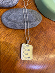 The Saved & remade necklace Porcelain by BUTIK KUBIK features a rectangular pendant of recycled porcelain with a line and dot design, hanging from a delicate silver-plated chain. It is shown resting on gray stone atop wood, with a green object in the background.