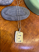 The Saved & remade necklace Porcelain by BUTIK KUBIK features a rectangular pendant of recycled porcelain with a line and dot design, hanging from a delicate silver-plated chain. It is shown resting on gray stone atop wood, with a green object in the background.