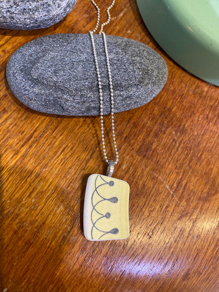 The Saved & remade necklace Porcelain by BUTIK KUBIK features a rectangular pendant of recycled porcelain with a line and dot design, hanging from a delicate silver-plated chain. It is shown resting on gray stone atop wood, with a green object in the background.