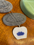 The Saved & remade necklace Porcelain by BUTIK KUBIK features a heart-shaped pendant of återbrukat porslin with blue flowers, resting on gray stones. A silverpläterad kedja and a green ceramic dish add contrast in the wooden background.