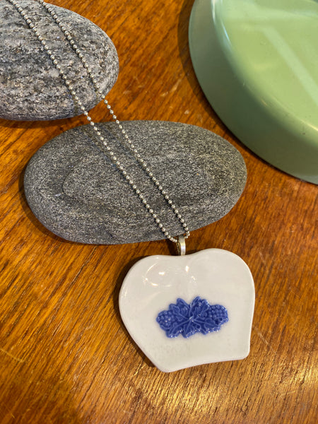 The Saved & remade necklace Porcelain by BUTIK KUBIK features a heart-shaped pendant of återbrukat porslin with blue flowers, resting on gray stones. A silverpläterad kedja and a green ceramic dish add contrast in the wooden background.