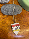 The Saved & Remade Necklace Porcelain by BUTIK KUBIK features a pendant of upcycled porcelain with a red, white, and yellow geometric design on a silver-plated chain, displayed on a gray stone over wood.