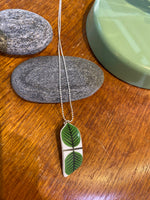 The Saved & Remade Necklace Porcelain by BUTIK KUBIK features a green leaf design pendant made from återbrukat porslin, shown resting on smooth gray stones atop wood, with a green dish partially visible.