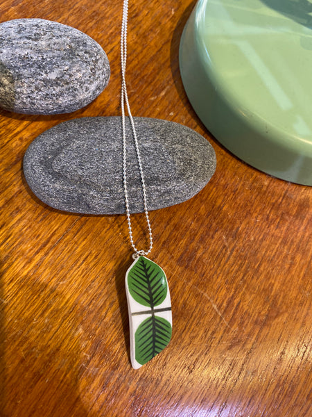The Saved & Remade Necklace Porcelain by BUTIK KUBIK features a green leaf design pendant made from återbrukat porslin, shown resting on smooth gray stones atop wood, with a green dish partially visible.