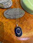 Saved & remade necklace Porcelain från BUTIK KUBIK: ett silverpläterat halsband med oval berlock av återbrukat porslin, dekorerad med mörkblå sten och vita cirklar. Smycket fotas mot grå sten på trä, med grönt inslag och fler stenar intill.