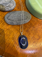 Saved & remade necklace Porcelain från BUTIK KUBIK: ett silverpläterat halsband med oval berlock av återbrukat porslin, dekorerad med mörkblå sten och vita cirklar. Smycket fotas mot grå sten på trä, med grönt inslag och fler stenar intill.