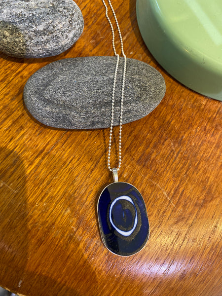 Saved & remade necklace Porcelain från BUTIK KUBIK: ett silverpläterat halsband med oval berlock av återbrukat porslin, dekorerad med mörkblå sten och vita cirklar. Smycket fotas mot grå sten på trä, med grönt inslag och fler stenar intill.