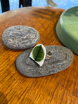 The Saved & remade ring porslin by BUTIK KUBIK is a silverplated ring featuring a white and green leaf-shaped piece of upcycled porcelain inspired by Berså Stig Lindberg, displayed on gray stones with a green accent in the background.