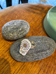 The Saved & remade ring porslin by BUTIK KUBIK is a silverplated ring featuring a white, oval ceramic top with delicate floral geometric design, crafted from recycled porcelain. Displayed on a smooth gray stone atop wood.