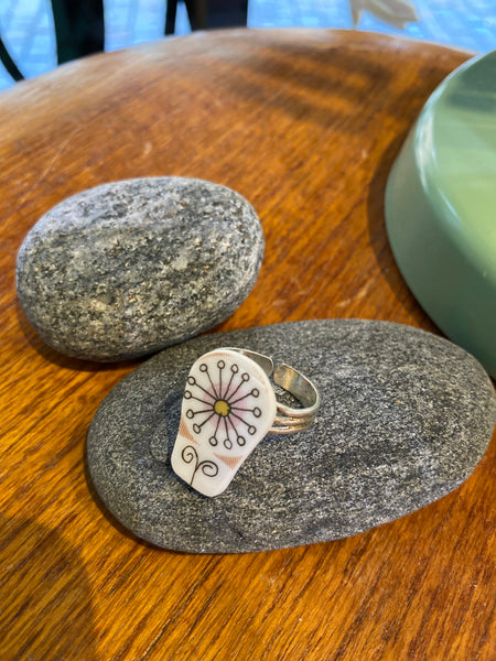 The Saved & remade ring porslin by BUTIK KUBIK is a silverplated ring featuring a white, oval ceramic top with delicate floral geometric design, crafted from recycled porcelain. Displayed on a smooth gray stone atop wood.