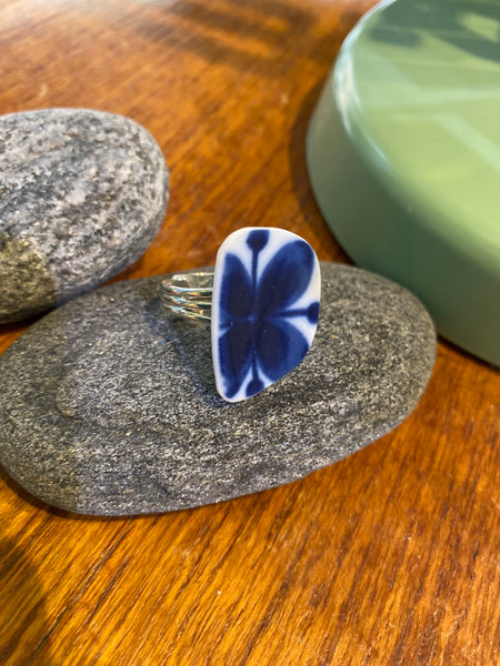 The Saved & remade ring porslin by BUTIK KUBIK is a silverplated ring with a rectangular blue and white Mon Amie ceramic floral piece, shown on a gray stone atop wood, with other stones and a green element nearby.