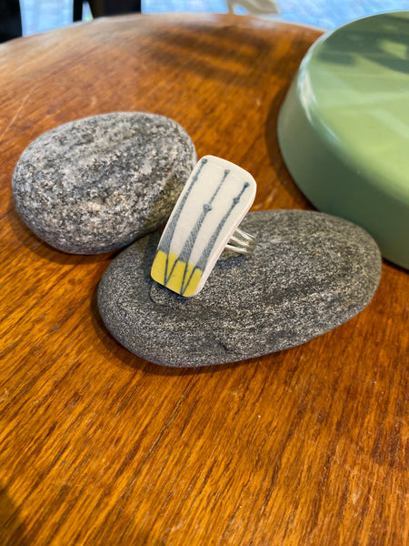 The Saved & remade ring porslin by BUTIK KUBIK is a rectangular, silver-plated ring made from upcycled porcelain featuring a white base, blue lines, and yellow tips, displayed on smooth gray stones atop wood beside a green object.
