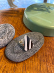 The Saved & Remade Ring Porslin by BUTIK KUBIK features a silver-plated band and a triangular black-and-white striped stone, displayed on a gray pebble beside a green ceramic container.
