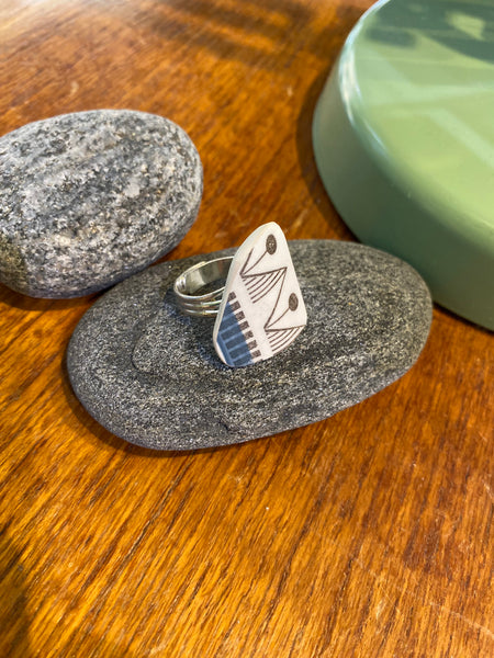 En Saved & remade ring porslin från BUTIK KUBIK står upprätt på en slät grå sten, med en till sten och ett grönt runt objekt intill på en träyta.