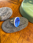 The Saved & remade ring porslin by BUTIK KUBIK features a round blue and white ceramic face with the GUSTAVSBERG PRUNUS inscription, silverplated band, displayed on smooth gray stones with a green accent nearby.