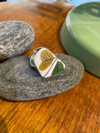 The Saved & remade ring porslin by BUTIK KUBIK is a silverplated ring featuring a green, yellow, and black leaf-patterned ceramic design from recycled porcelain, displayed on gray stones over wood with a green object in the background.
