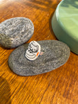 The BUTIK KUBIK Saved & remade ring porslin features a white, oval upcycled porcelain face with a black cat line drawing, shown on a smooth gray stone next to another stone and a green item on wood.