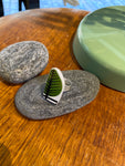 The Saved & Remade Ring Porslin by BUTIK KUBIK, silver-plated and featuring a green leaf Berså Stig Lindberg design, stands upright on a smooth gray stone atop a wooden surface with another stone and a pale green item nearby.