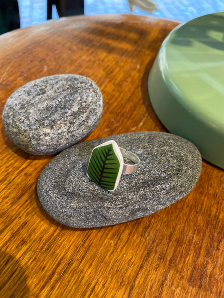 The Saved & remade ring porslin by BUTIK KUBIK, featuring a green leaf motif, sits on a smooth gray stone with decorative stones and a round green accent in the background.