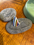 The Saved & remade ring porslin by BUTIK KUBIK features a silverplated band with a triangular top made from recycled porcelain, showcasing blue, green, red, and black stripes—a distinctive accessory displayed on gray stones beside a green accent.