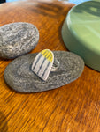The Saved & remade ring porslin by BUTIK KUBIK is a silverplated ring with a flat, triangular top made from upcycled porcelain featuring yellow and black lines, shown resting on a gray stone with other objects on a wooden surface.