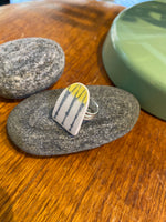 The Saved & remade ring porslin by BUTIK KUBIK is a silverplated ring with a flat, triangular top made from upcycled porcelain featuring yellow and black lines, shown resting on a gray stone with other objects on a wooden surface.