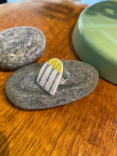 The Saved & remade ring porslin by BUTIK KUBIK is a silverplated ring with a flat, triangular top made from upcycled porcelain featuring yellow and black lines, shown resting on a gray stone with other objects on a wooden surface.