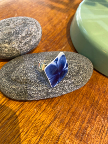 The Saved & remade ring porslin by BUTIK KUBIK—a silverplated ring featuring blue-and-white upcycled porcelain—rests on a smooth gray stone, with another stone and a green ceramic object in the background on wood.