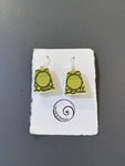 Saved & remade earrings by BUTIK KUBIK: green rectangular återbrukat porslin with black patterns, displayed on a white textured card with the Saved and Remade spiral logo and text, set against a gray background.