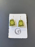 Saved & remade earrings by BUTIK KUBIK: green rectangular återbrukat porslin with black patterns, displayed on a white textured card with the Saved and Remade spiral logo and text, set against a gray background.