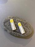 A pair of rectangular Saved & remade earrings by BUTIK KUBIK, crafted from återbrukat porslin with yellow and blue abstract designs and gold hooks, rest on an oval gray stone against a gray background.