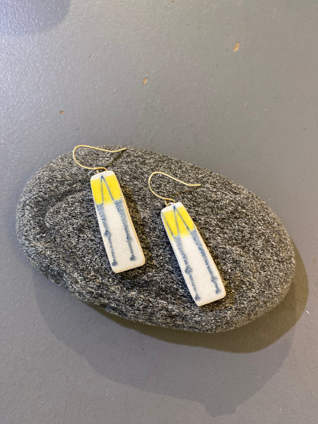 A pair of rectangular Saved & remade earrings by BUTIK KUBIK, crafted from återbrukat porslin with yellow and blue abstract designs and gold hooks, rest on an oval gray stone against a gray background.