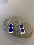 A pair of BUTIK KUBIKs Saved & remade earrings—silverplated, with rectangular white bases of upcycled porcelain and two dark blue circular accents—shown on an oval gray stone against a gray backdrop.
