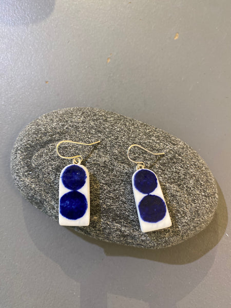 A pair of BUTIK KUBIKs Saved & remade earrings—silverplated, with rectangular white bases of upcycled porcelain and two dark blue circular accents—shown on an oval gray stone against a gray backdrop.