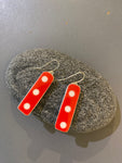 The Saved & remade earrings by BUTIK KUBIK are two rectangular red pieces with three white dots each, made from återbrukat porslin and displayed on a smooth gray stone against a gray background.