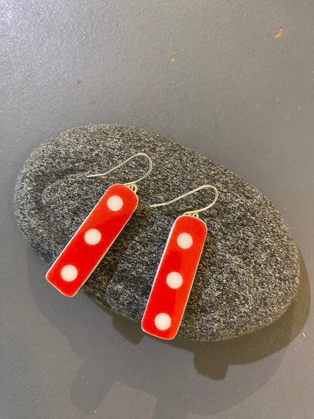 The Saved & remade earrings by BUTIK KUBIK are two rectangular red pieces with three white dots each, made from återbrukat porslin and displayed on a smooth gray stone against a gray background.