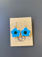Saved & remade earrings by BUTIK KUBIK: Blue and white flower-shaped pendants made from recycled porcelain, with dark centers and silver-plated hooks. Displayed on a brown card with spiral logo, placed on a gray surface.