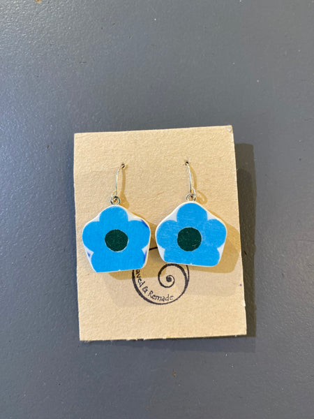 Saved & remade earrings by BUTIK KUBIK: Blue and white flower-shaped pendants made from recycled porcelain, with dark centers and silver-plated hooks. Displayed on a brown card with spiral logo, placed on a gray surface.