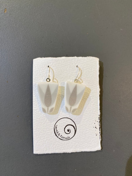 A pair of geometric white Saved & remade earrings by BUTIK KUBIK, featuring gray leaf designs on upcycled porcelain, are shown on a textured card with a spiral logo and “Seeks & Remade”; background is gray.