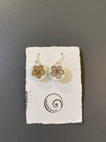 A pair of Saved & remade earrings by BUTIK KUBIK—white silverplated with brown floral designs, made from recycled porcelain, shown on a textured white card with spiral logo and Saved and Remade text, set against a gray background.